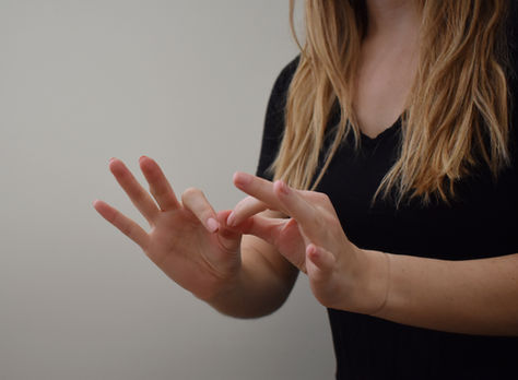 A person with long hair signs in American Sign Language against a plain background, wearing a black shirt. Hands are in focus.
