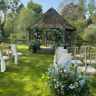 blue and white spray of flowers in the foreground, with chairs draped in white and the lake summerhouse behind 