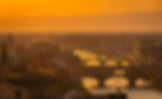 Sunset over Florence with orange hues, featuring the Ponte Vecchio and cityscape. Boats on the Arno River, creating a serene atmosphere.