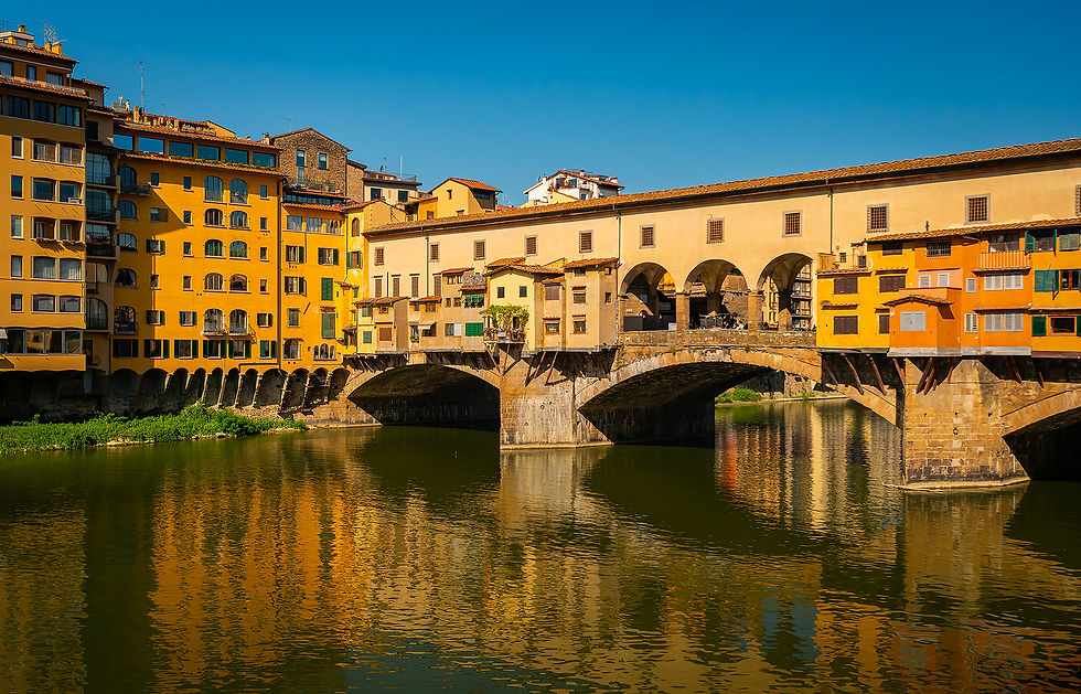 Historic bridge over a river with colorful buildings and arches, reflecting in calm water under a clear blue sky. Peaceful and vibrant scene.