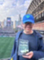 Smiling person in a blue cap holds Phantom Algebra book at a crowded stadium. Lumen Field signs and city skyline in background.