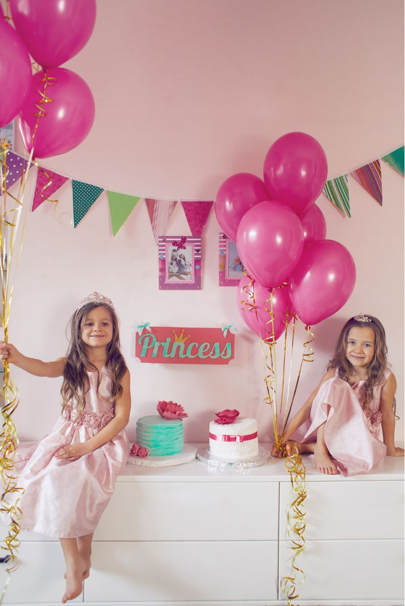 Two children in pink dresses sit by cakes and pink balloons in a decorated party room with "Princess" sign.