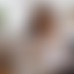 Smiling woman in beige blazer at desk, reviewing papers, holding glasses. Laptop, phone, and plants in bright modern office setting.