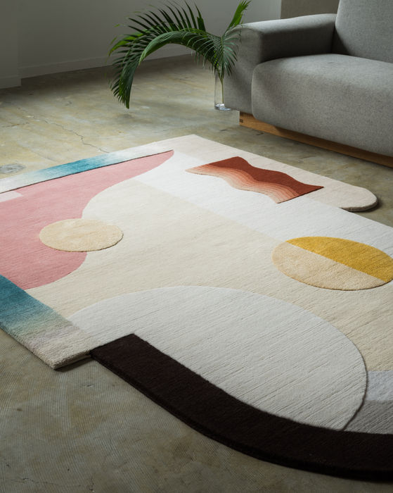 A detail of Jensin Okunishi Studio handmade rug. The abstract rug is composed of various geometric shapes and soft colors. The edges are also curved and asymmetrical. A palm leaf sits next to a grey couch on the cement floor. 
