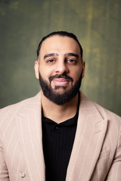 A portrait by poet Taher Adel. He is smiling and wears a black t-shirt under a beige suit, against a green background.