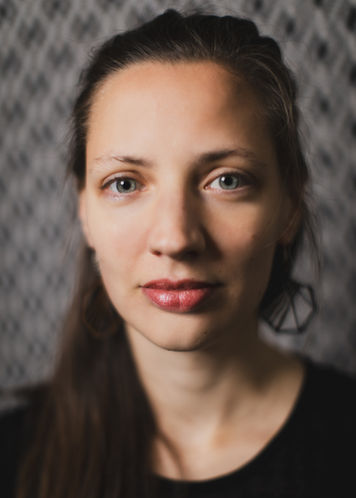 A headshot of Marie Schreer against a sightly blurry chequered background. She looks friendly yet deep in thought.