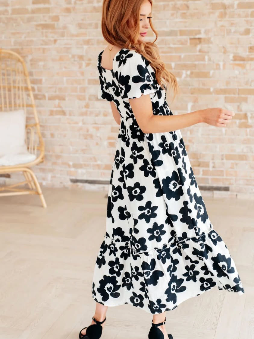 Thumbnail: Woman wearing floral dress, posing. Smiling, near a wicker chair and wall.