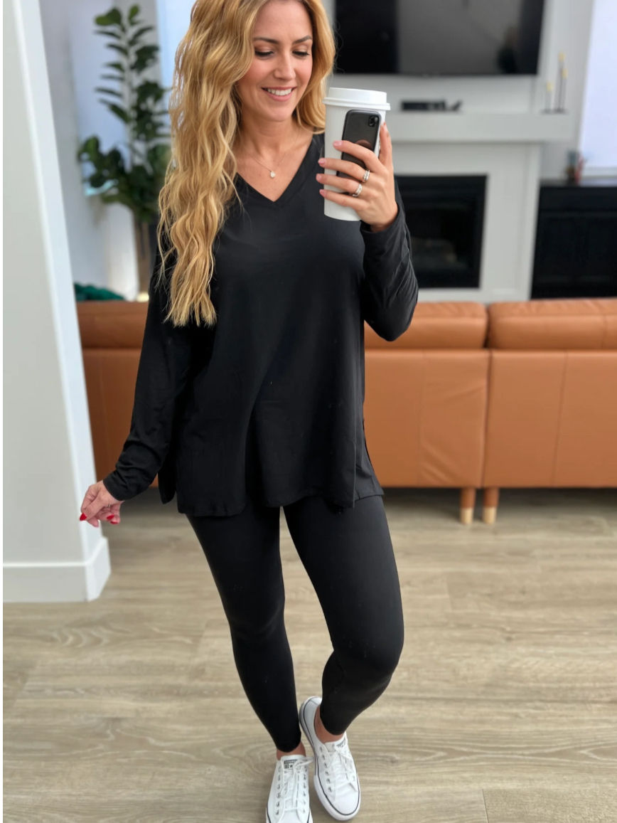 Woman in black outfit holding phone, smiling in front of couch.