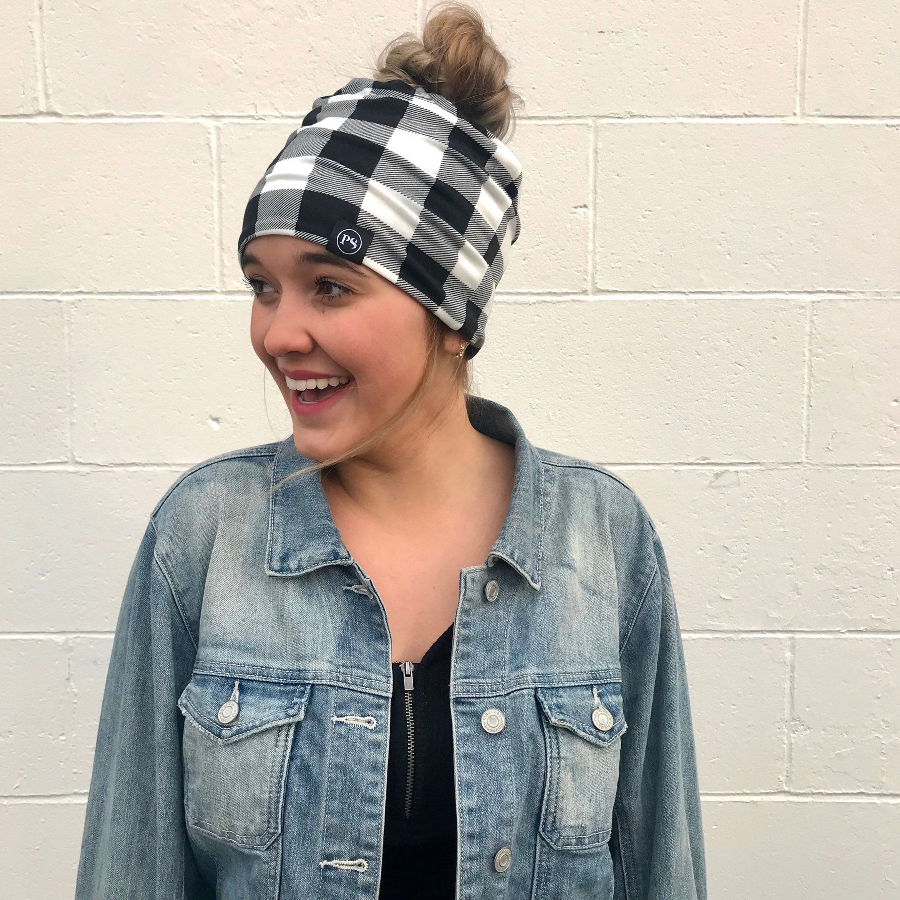 Smiling woman wearing plaid beanie and denim jacket against a white brick wall