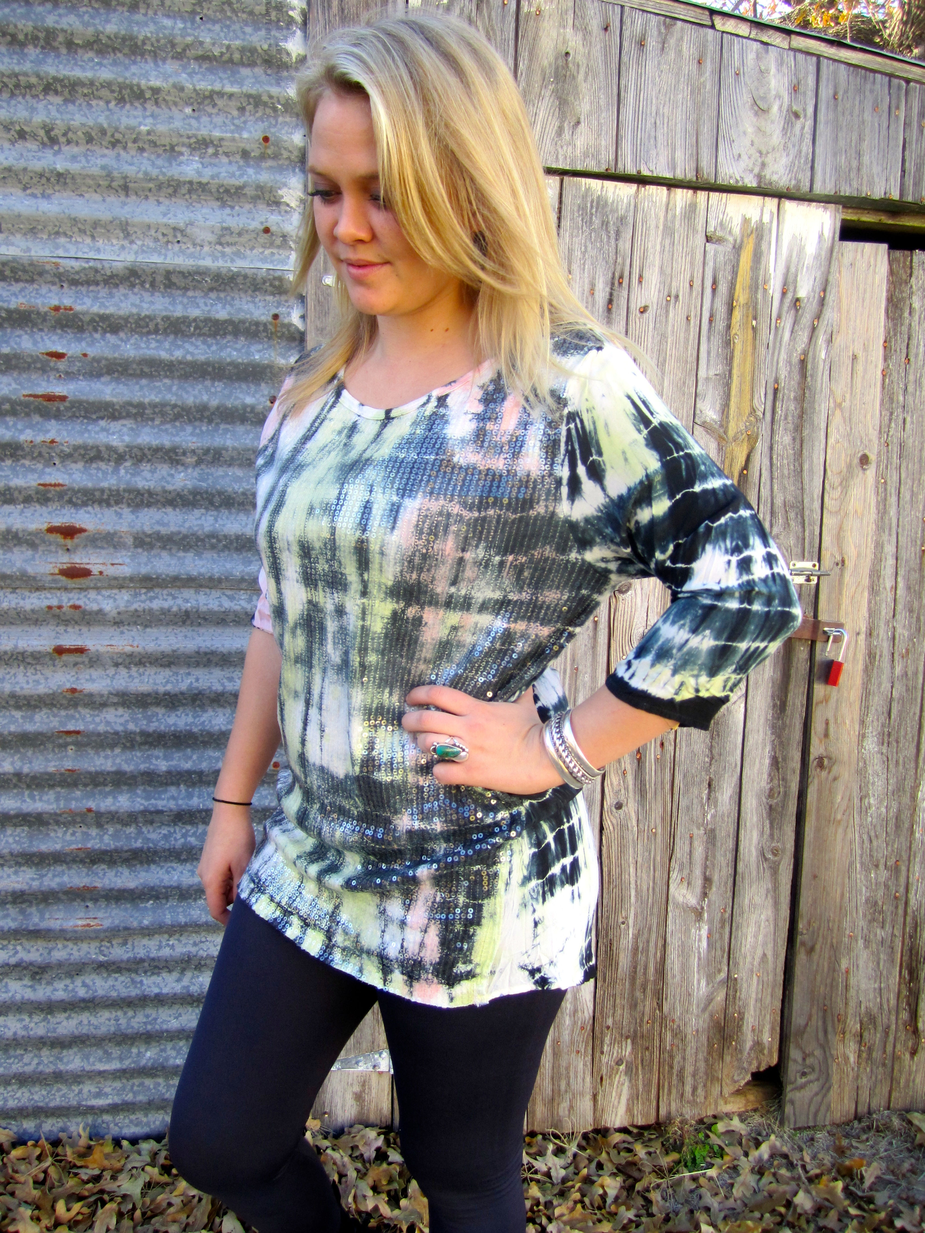 Woman wearing a tie-dye shirt standing near a wooden door, smiling slightly.