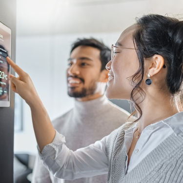 Woman adjusting smart home control panel while man watches smiling. Screen displays temperature 23°C. Modern, bright indoor setting.
