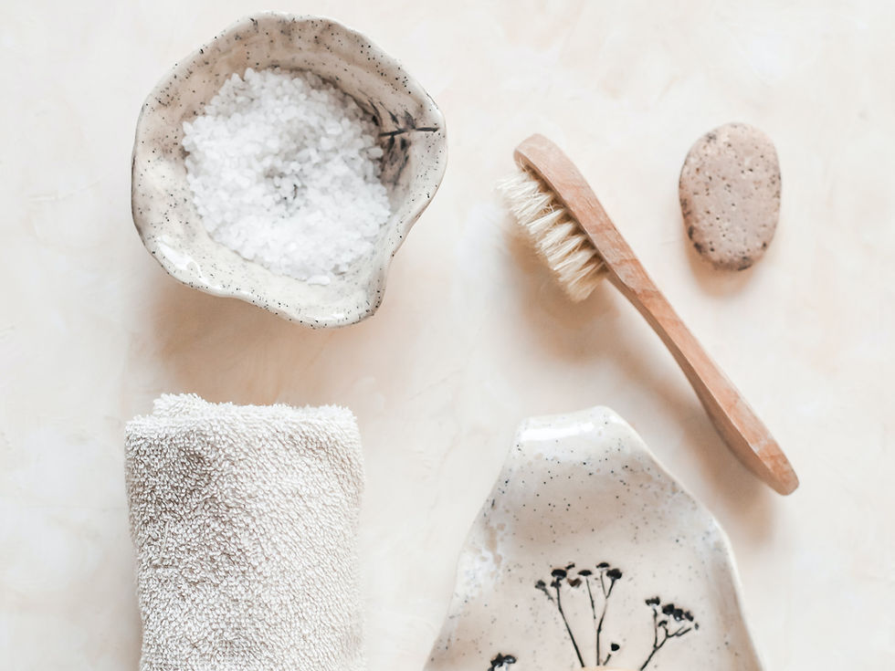 A spa-inspired salt scrub featuring a textured ceramic bowl filled with coarse salt, accompanied by a wooden brush, a pumice stone, and a soft towel on a light background.
