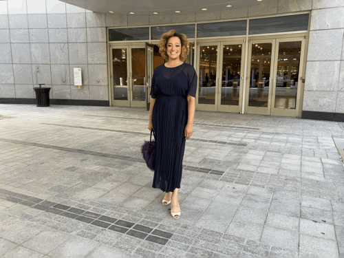 Woman in navy dress smiles, holds purple bag outside an office building with glass doors. Gray stone facade; mood is cheerful.