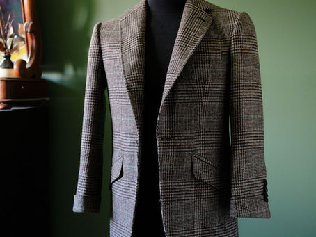 A beautifully tailored tweed sport coat with a subtle plaid pattern on a black mannequin, set against a deep olive green wall. The jacket features notched lapels, two flapped pockets, and a clean, structured silhouette. A dark wooden mirror or vanity is partially visible in the background on the left.