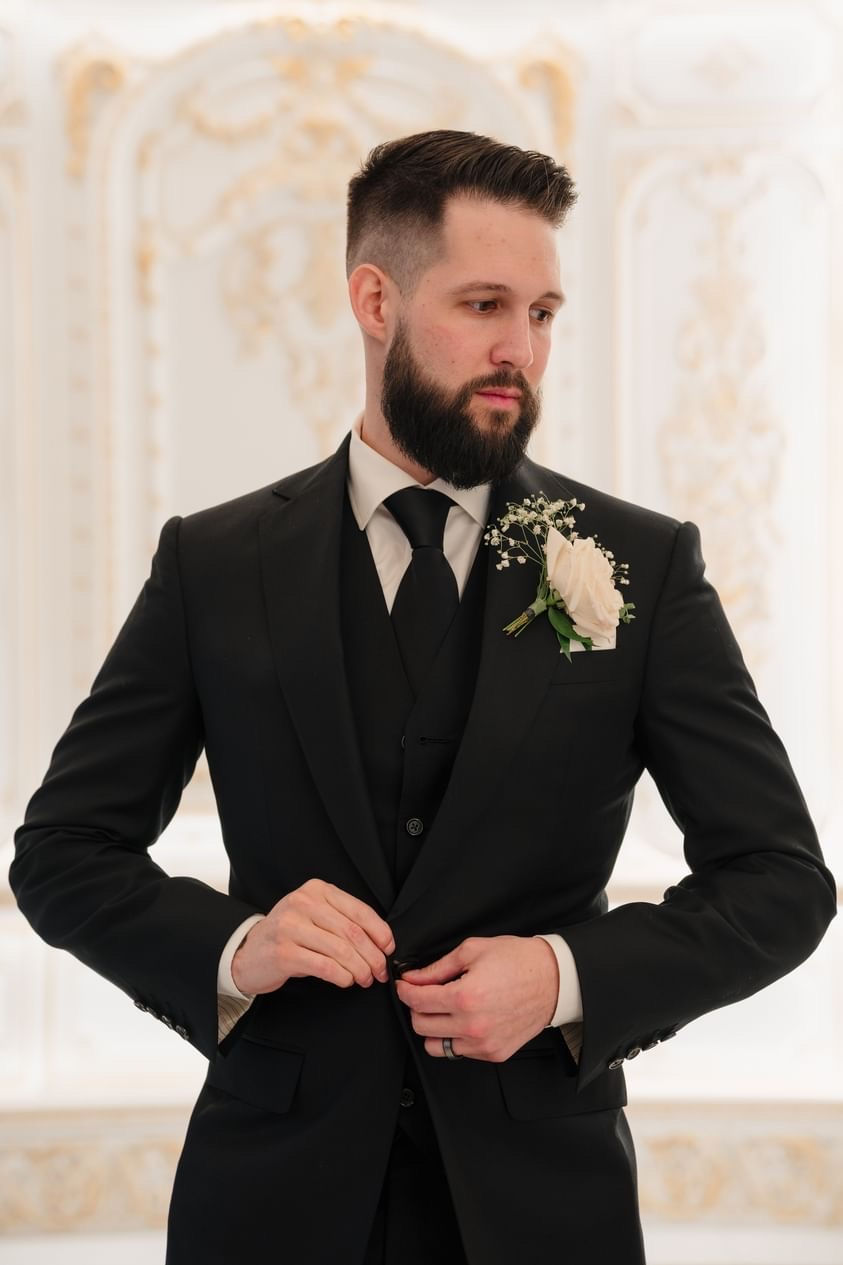 Groom wearing a black bespoke wedding suit with boutonnière, adjusting jacket in an elegant setting.