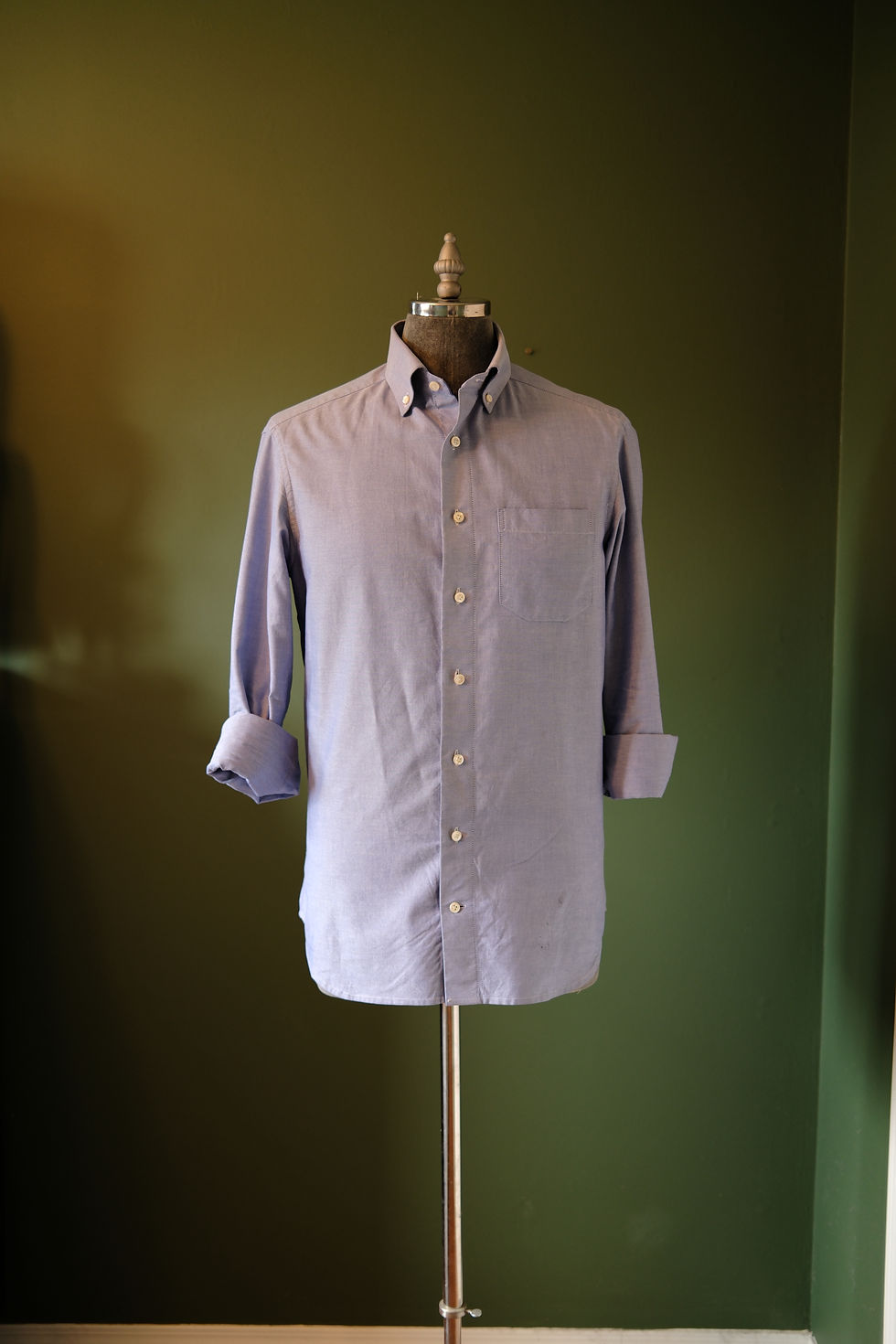 A light blue, long-sleeved dress shirt with a button-down collar and a single chest pocket, displayed on a dark-colored mannequin against a deep olive green wall. The sleeves are rolled up to the elbows, revealing the cuffs.