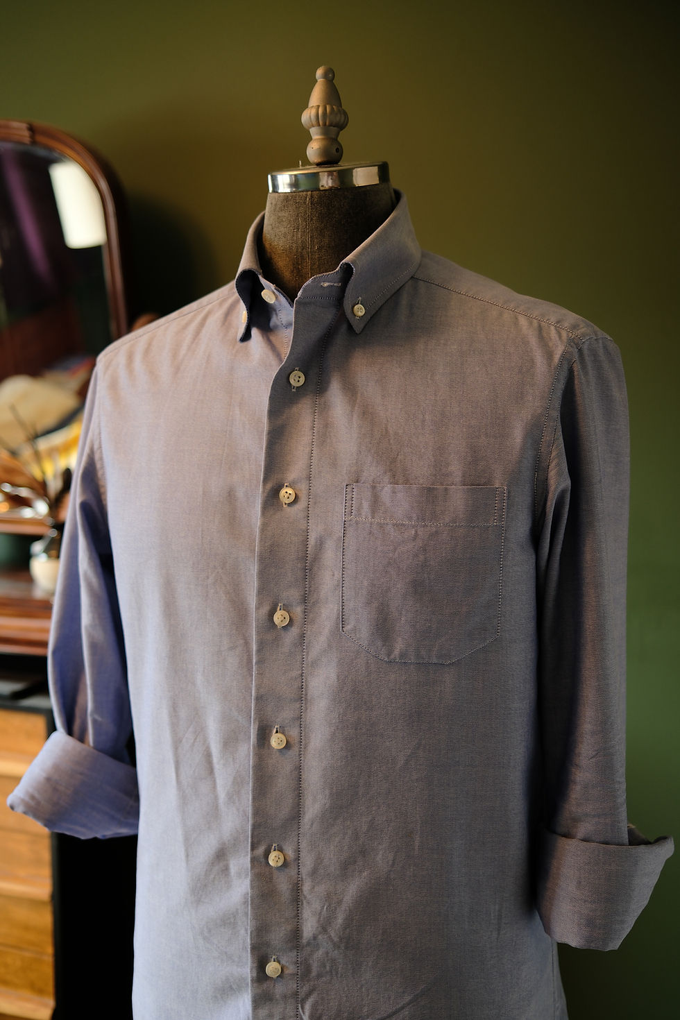 A close-up, angled shot of a light blue long-sleeved dress shirt with a button-down collar and a chest pocket, displayed on a dark mannequin. The shirt's fabric texture and stitching details are visible, and the sleeves are rolled. A blurred reflection of a mirror and a wooden furniture piece are in the background.