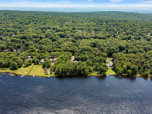 An aerial view of a lakefront residential property in the Poconos, featuring a spacious waterfront lot with a private dock, manicured lawn, and mature trees. The home sits along a peaceful shoreline surrounded by dense forest, with neighboring lakefront properties and expansive woodland stretching to the horizon under a clear blue sky.