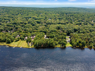 An aerial view of a lakefront residential property in the Poconos, featuring a spacious waterfront lot with a private dock, manicured lawn, and mature trees. The home sits along a peaceful shoreline surrounded by dense forest, with neighboring lakefront properties and expansive woodland stretching to the horizon under a clear blue sky.