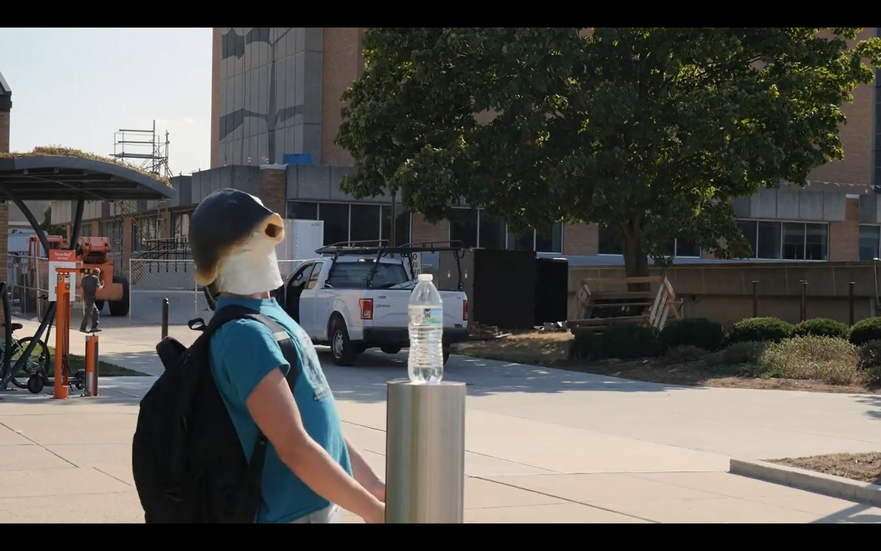 Man with a fish mask looking at a water bottle, outside.