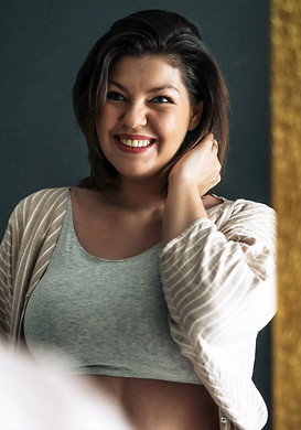 A woman with short brown hair, wearing a light gray crop top and a striped cardigan, smiles while touching her neck in front of a mirror, perhaps reflecting after her bariatric mental health evaluation.