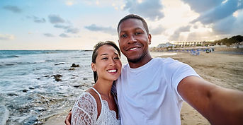 A couple stands on a beach taking a selfie, with the ocean and cloudy sky in the background, capturing a moment of togetherness as they focus on conflict resolution and strengthening their bond.