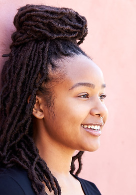 Young black Woman with long, styled locs and a nose ring smiling while standing against a light pink wall, radiating confidence and well-being—reflecting the positive impact of online therapy on her mental health.