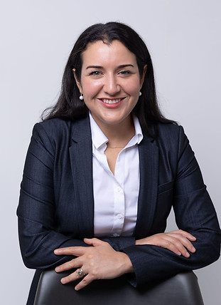 Alicia Ciliezar with long dark hair wearing a navy blazer and white shirt, smiling and sitting with arms crossed on the back of a chair, representing medication management against a plain light background.