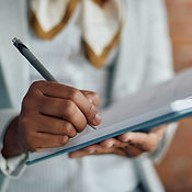 A person holds a pen and writes on a notepad, with glasses and papers visible in the blurred background, perhaps jotting down notes from a self-pay counseling or affordable therapy options session.