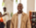 A person in a tan jacket and white shirt stands indoors, smiling; other people and plants are visible in the blurred background, suggesting a welcoming space where affordable therapy options or sliding scale therapy may be available.