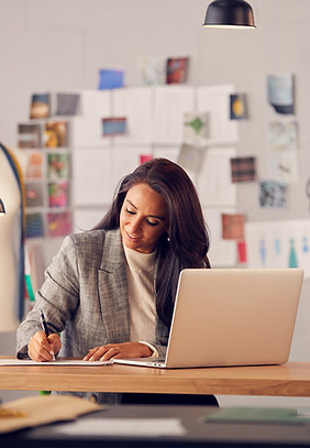 A latina woman in a gray blazer writes on paper while sitting at a desk with a laptop in a modern office setting, reflecting on stress management counseling techniques.