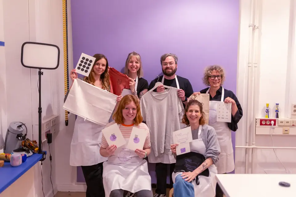 Siebdruck-Workshop Hamburg: Gruppe von Teilnehmenden präsentiert ihre selbstgedruckten T-Shirts, Taschen und Karten vor einer lila Wand.
