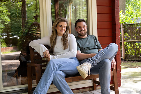 De hosts Emmy en Gertjan glimlachend op de foto op een bankje op de veranda van Boshuisje de Kempen