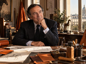 Jean-Louis Dumas-inspired portrait in a Parisian leather atelier surrounded by craftsmanship tools, leather materials, sketches and a structured luxury handbag.