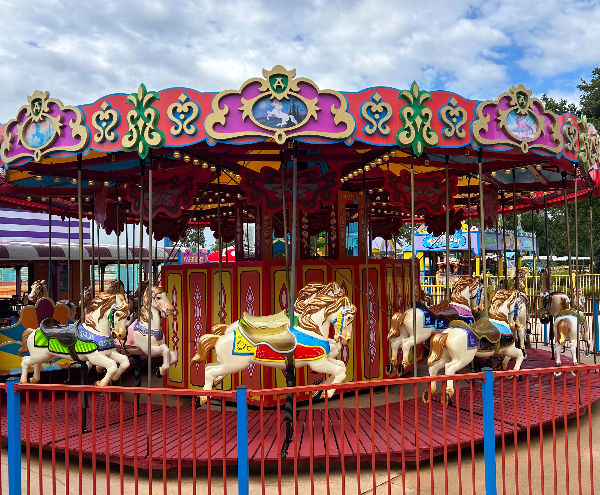 Colourful carousel with ornate patterns and horse figures in a park under a cloudy sky. The atmosphere is playful, surrounded by a red fence.