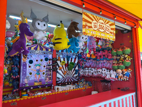 Carnival booth with colourful stuffed animals, including a dragon and unicorn. Price sign: $8, $50, $20. Vibrant, playful atmosphere.