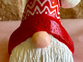 A cute gnome figurine with a red hat featuring white patterns and a fluffy white beard, set against a brick wall background.