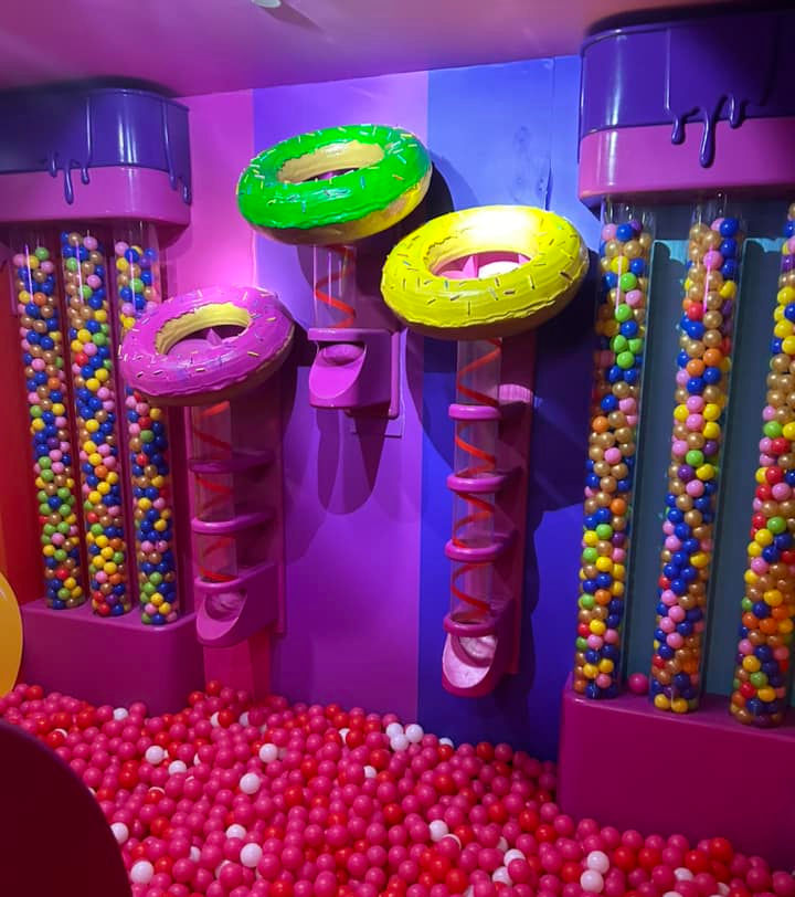 Bright play area with donut-themed slides, colorful ball pit, and candy ball tubes. Walls in pink and purple, creating a playful mood.