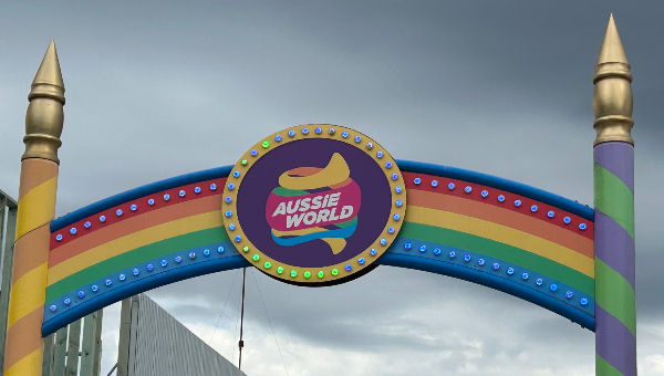 Rainbow-colored arch with "Aussie World" sign, lit by colorful bulbs, set against a cloudy sky. Vertical striped pillars flank the arch.