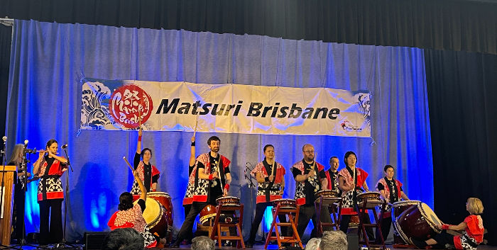 People in traditional attire perform Taiko drumming on stage at Matsuri Brisbane. A banner and blue lighting create an energetic scene.