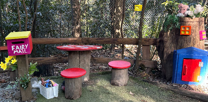Colourful fairy-themed play area with red mushroom table and stools, a pink "Fairy Mail" box, and toys in a basket against a forest backdrop.