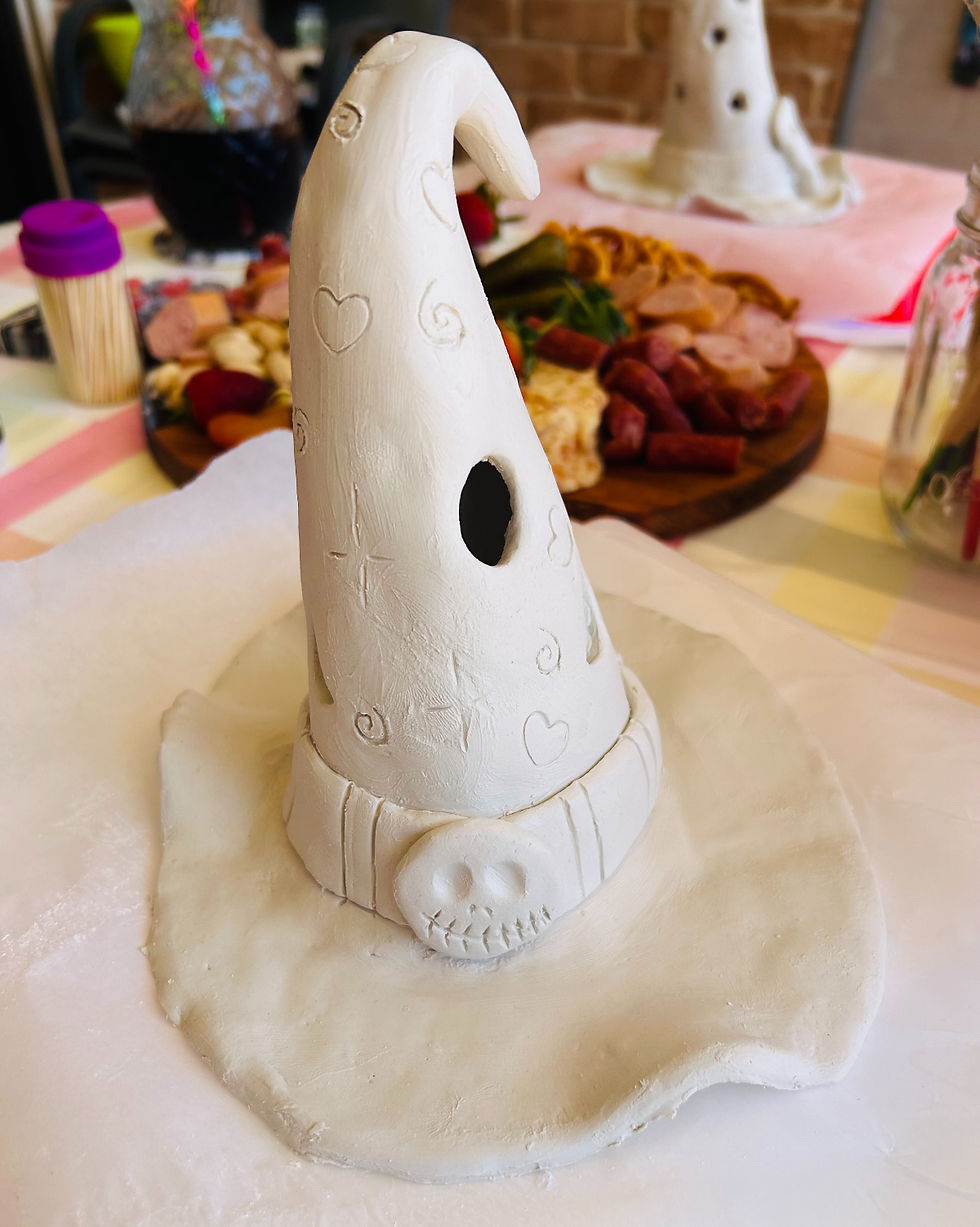White clay witch hat with heart and swirl designs on a table. Skull decoration on brim. Background of food platter and jar. Cozy setting.