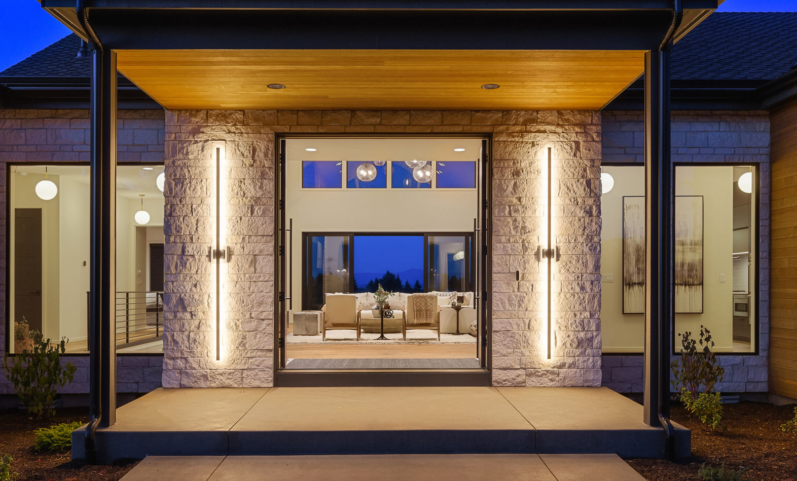 Entrance to stunning luxury home at twilight. Double doors are open, inviting the viewer to look through the entrance flanked by textured stone and vertical sconce lights through the great room and to the sliding glass doors leading to a deck with a viewing platform.
