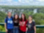 A family of 4 pose for a camera with a background of 100s of waterfalls and greenery. They all smile, gently embracing one another. The mom and dad lovingly sandwich their 2 grown adult daughters. The shorter sister has a Giant Cape nevus covering her neck and chest. 