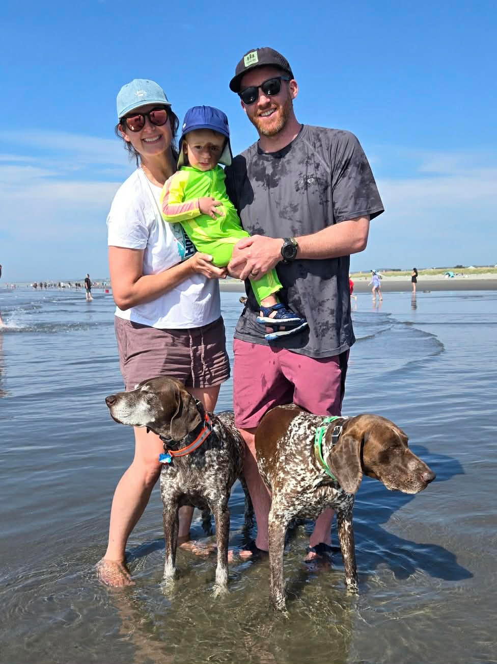 A family of 3 stands in the Oregon waters with their 2 speckled dogs. Wearing smiles, they hold their son, dressed in a lime green full body swimsuit. All 3 are practicing sun safety by wearing sunglasses,