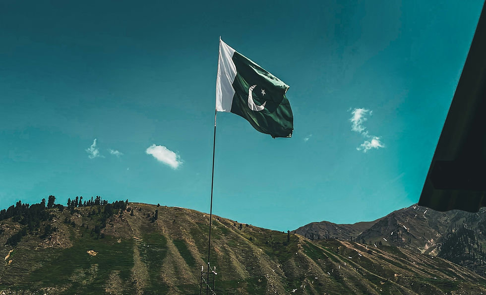 Pakistan flag waving on a tall pole above green mountain hills under a bright blue sky with small clouds.