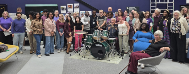 Large group smiles around a green Yamaha drum kit in a purple-walled room, posing for a group photo.