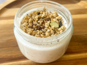 A glass jar filled with overnight oats made with grated peach, yogurt, and milk, topped with muesli and seeds, served on a wooden board in soft morning light.