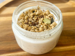 A glass jar filled with overnight oats made with grated peach, yogurt, and milk, topped with muesli and seeds, served on a wooden board in soft morning light.