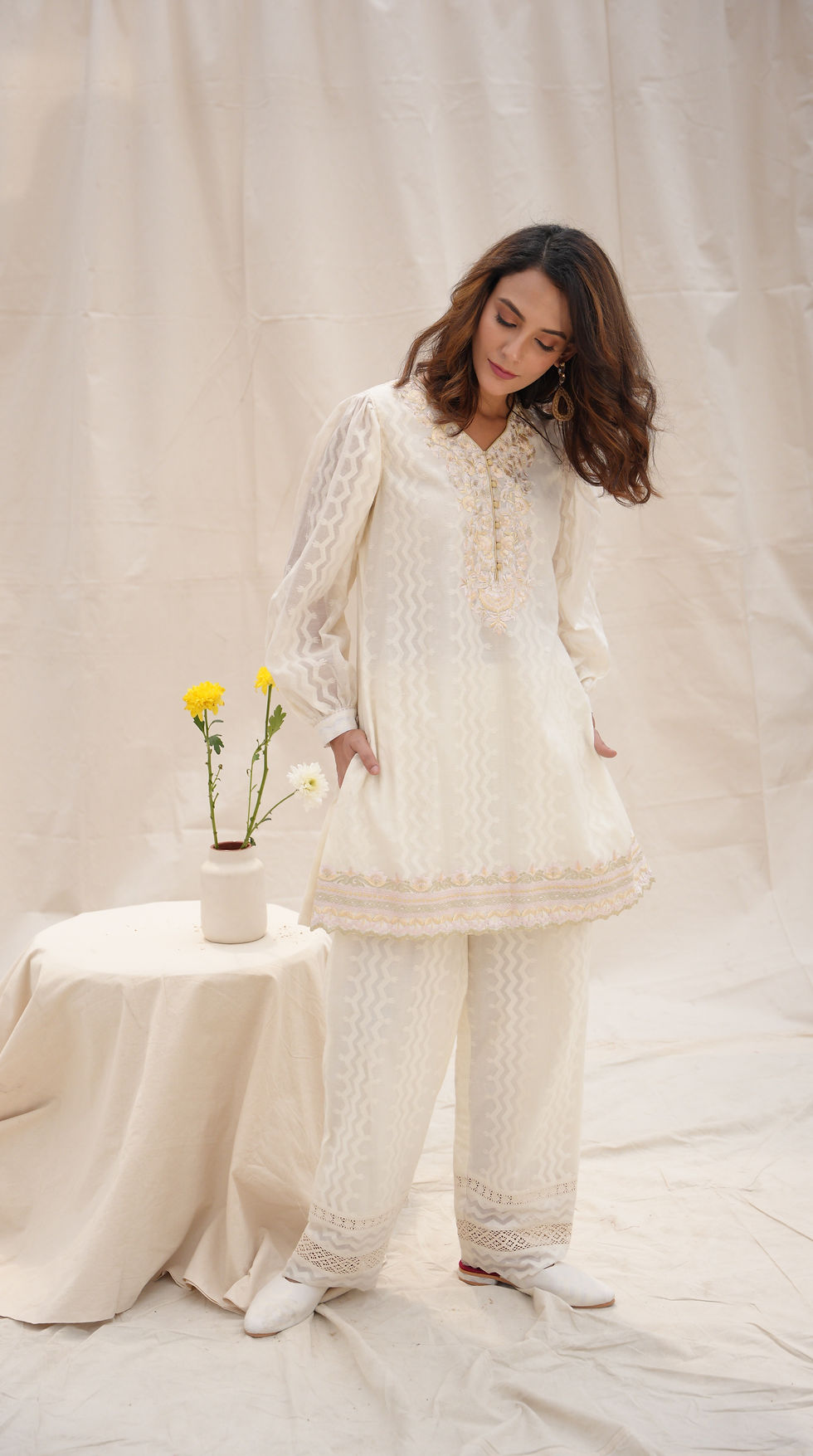 Thumbnail: A woman wearing an elegant cream embroidered giza cotton outfit set with hands in pockets. A small table with yellow flowers is beside her, set against a beige backdrop.